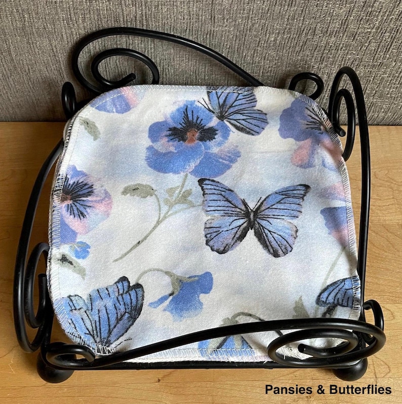 May include: A black metal napkin holder with a square base and scrolled wire corners, containing a square fabric napkin featuring a light blue background with a pattern of blue pansy flowers and blue butterflies, labeled Pansies & Butterflies.