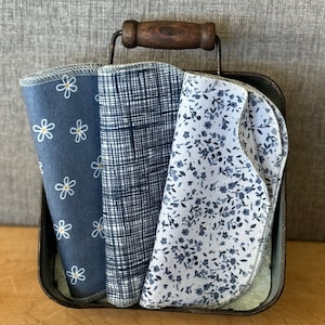 May include: Three folded fabric cloths with rounded edges and serged seams, featuring blue and white patterns including white daisies, a dark blue grid, and small blue floral prints, all contained within a dark metal bin with a wooden handle.