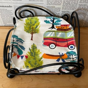 May include: A stack of white fabric squares with a colorful pattern of trees, a camper van, and a canoe. The fabric squares are folded and stacked on a black metal stand.