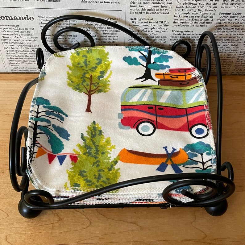 May include: A stack of white fabric squares with a colorful pattern of trees, a camper van, and a canoe. The fabric squares are folded and stacked on a black metal stand.