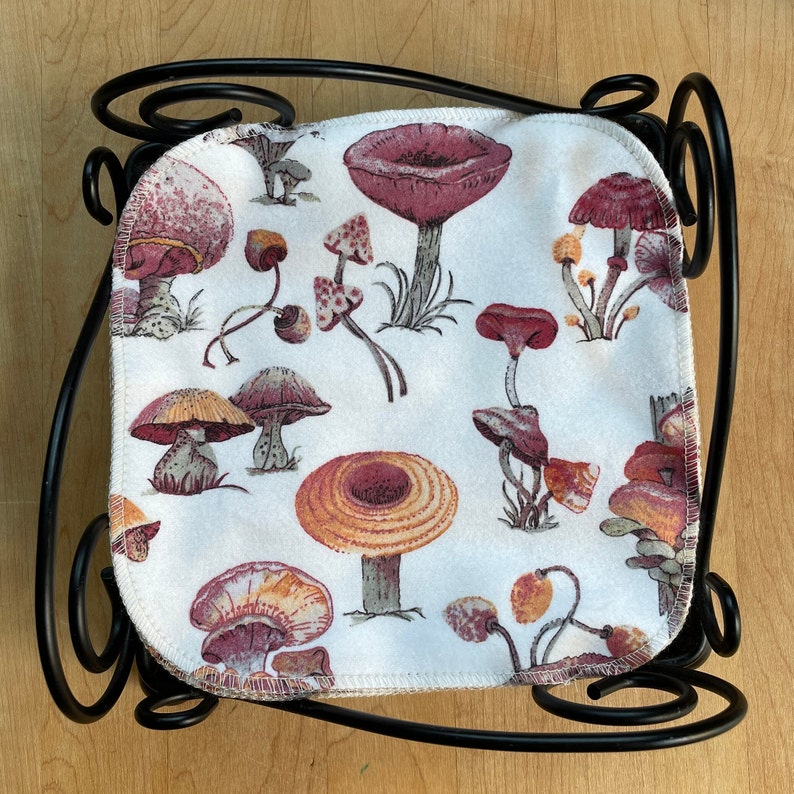 May include: A white fabric coaster with a black metal frame features a repeating pattern of red and brown mushrooms. The mushrooms are stylized and have a vintage look.