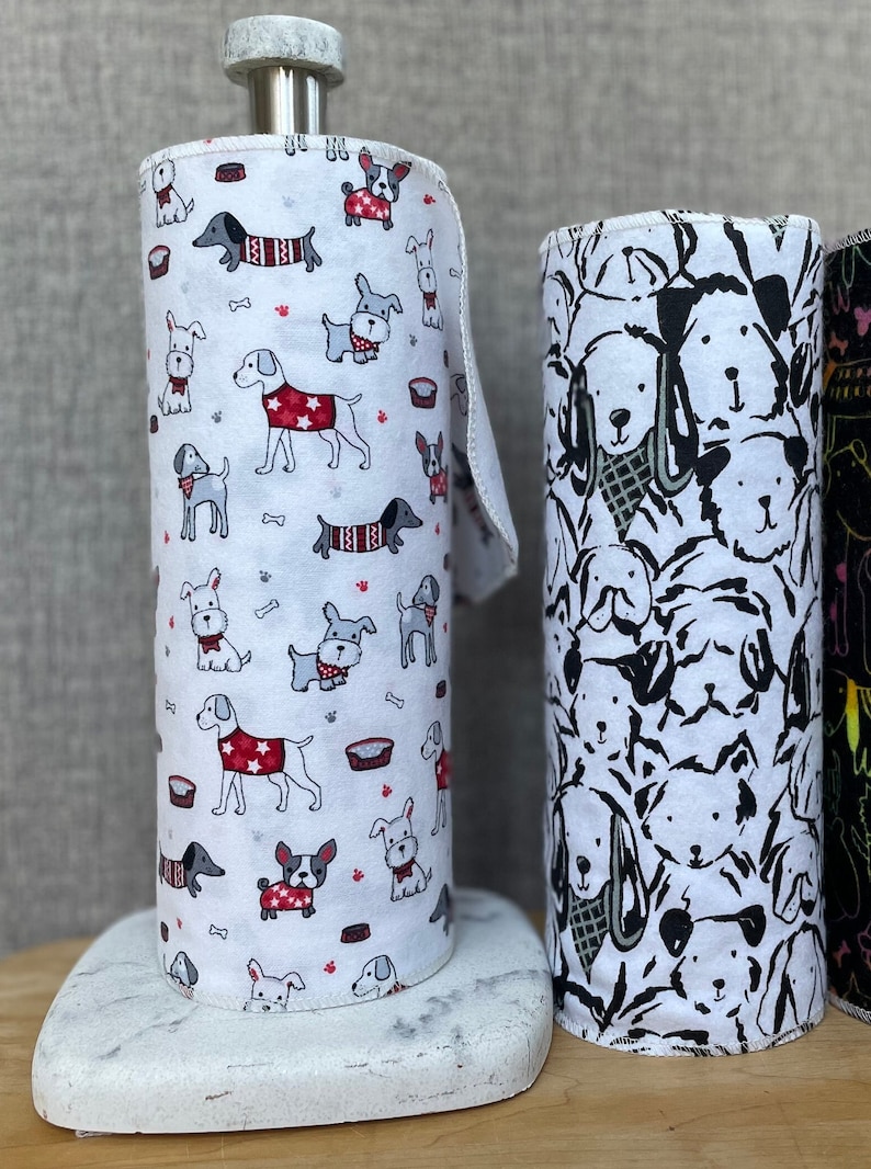 K&ouml;nnte beinhalten: Zwei Rollen wei&szlig;er Papierhandt&uuml;cher mit verschiedenen Hunde-Designs. Eine Rolle hat einen wei&szlig;en Hintergrund mit bunten Hunden, die Pullover und H&uuml;te tragen. Die andere Rolle hat ein Schwarzwei&szlig;-Design mit Hunden in verschiedenen Posen.