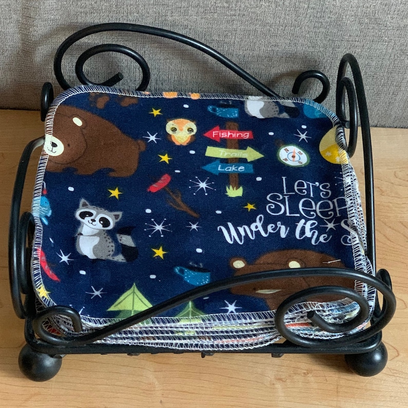 May include: A stack of blue fabric squares with a white border and a woodland animal print. The fabric squares are in a black metal holder. The fabric squares have a variety of woodland animals, including bears, owls, raccoons, and stars. The fabric squares also have the text "Let's Sleep Under the Stars" printed on them.