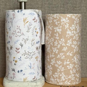 May include: Two cylindrical rolls of fabric featuring botanical prints. The left roll is white with a multi-colored floral pattern, while the right roll is tan with a white floral pattern. The left roll sits on a metal paper towel holder.
