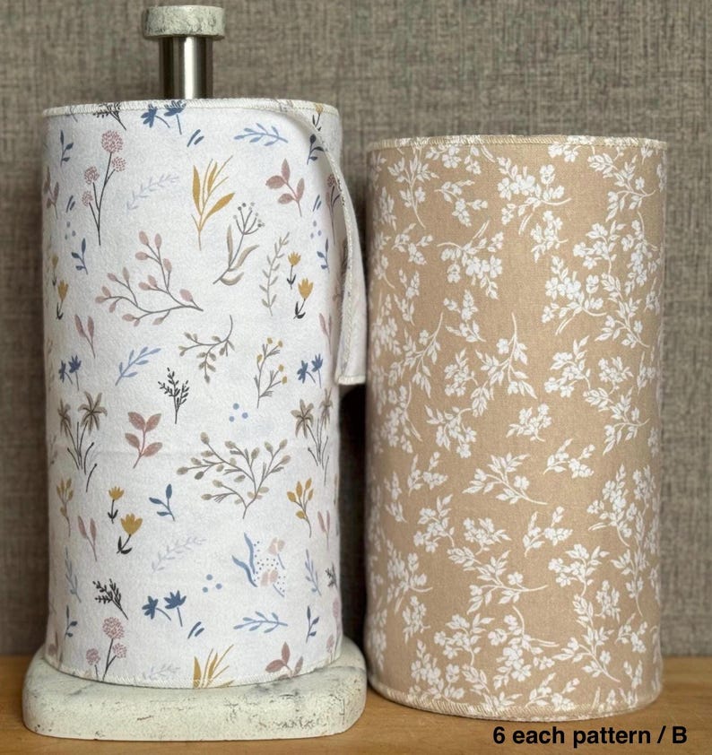 May include: Two cylindrical rolls of fabric featuring botanical prints. The left roll is white with a multi-colored floral pattern, while the right roll is tan with a white floral pattern. The left roll sits on a metal paper towel holder.