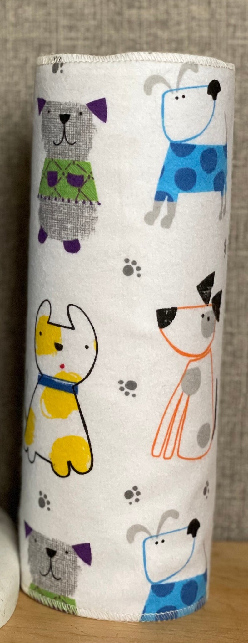 K&ouml;nnte beinhalten: Wei&szlig;er Papierhandtuchhalter mit einem bunten Muster aus Hunden. Die Hunde sind grau, gelb, orange und blau mit schwarzen Flecken. Der Hintergrund ist wei&szlig; mit grauen Pfotenabdr&uuml;cken.