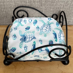 Pode incluir: Uma pilha de panos de tecido quadrados brancos com uma estampa de tartarugas marinhas e peixes em azul e turquesa, mantida dentro de um porta-guardanapos de metal preto com volutas decorativas e quatro pequenos p&eacute;s esf&eacute;ricos.
