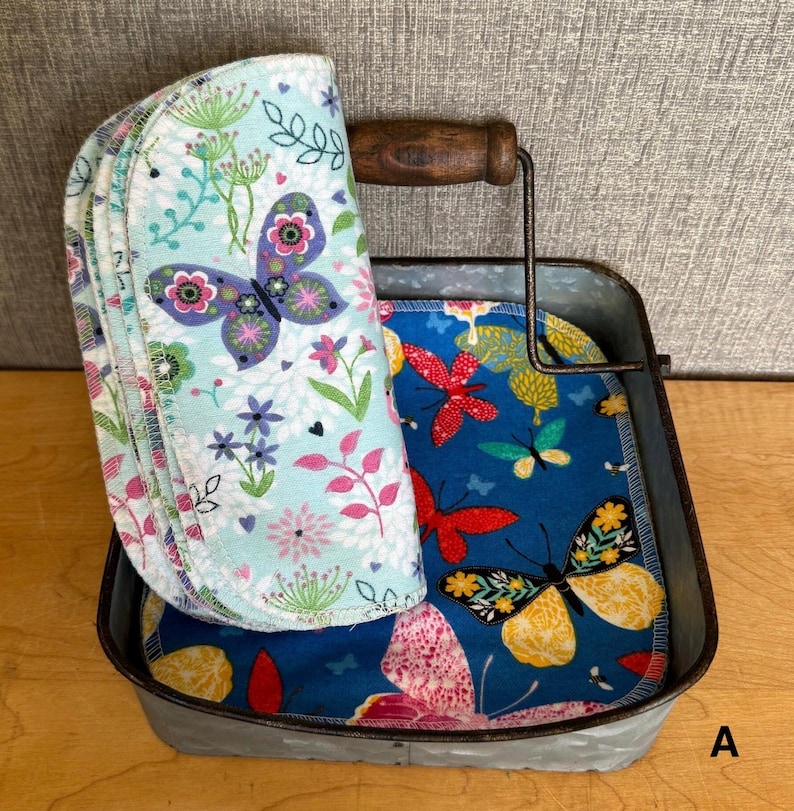 May include: A galvanized metal tray with a wooden handle containing several round fabric cloths. One cloth features a light blue background with colorful butterflies and floral patterns, while others inside the tray display a dark blue background with red, yellow, and orange butterflies.