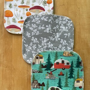 May include: Three square cotton dishcloths with different patterns. The top dishcloth has a white background with orange and red mushrooms. The middle dishcloth has a gray background with white floral designs. The bottom dishcloth has a teal background with a camping theme featuring red and green camper vans, trees, and animals.