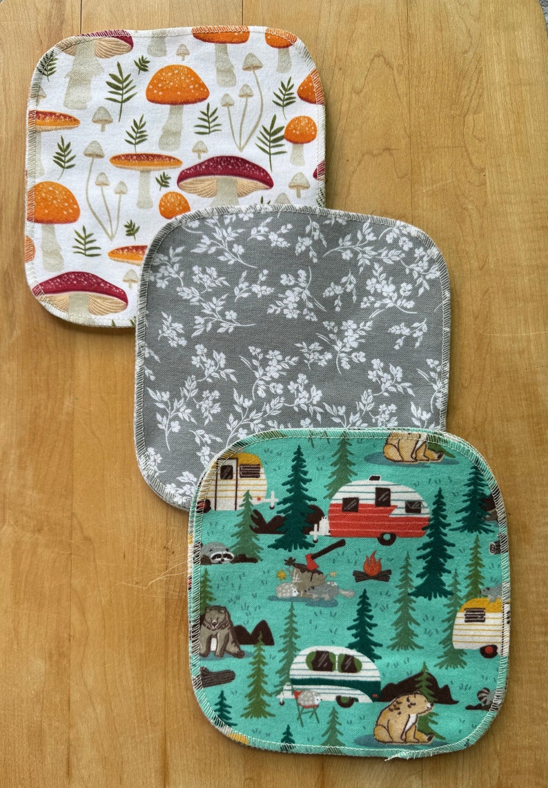 May include: Three square cotton dishcloths with different patterns. The top dishcloth has a white background with orange and red mushrooms. The middle dishcloth has a gray background with white floral designs. The bottom dishcloth has a teal background with a camping theme featuring red and green camper vans, trees, and animals.