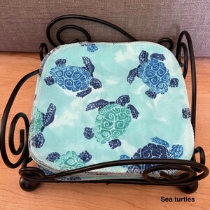 Pode incluir: Uma pilha de panos de tecido quadrados com um fundo azul claro e um padr&atilde;o repetitivo de tartarugas marinhas em azul escuro e azul-petr&oacute;leo, mantidos num suporte de arame de metal preto com detalhes decorativos em espiral nos cantos.