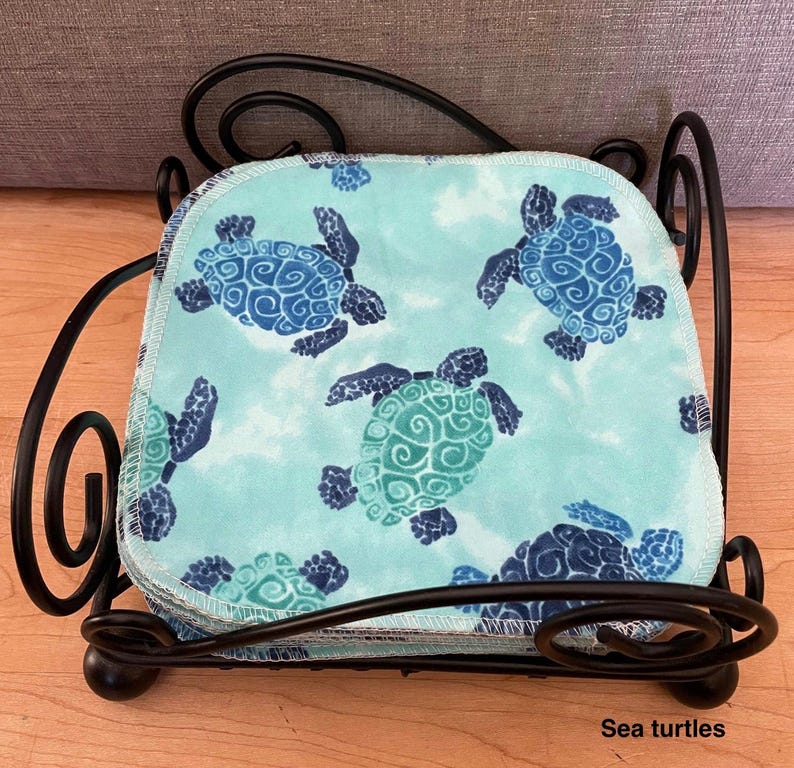 Pode incluir: Uma pilha de panos de tecido quadrados com um fundo azul claro e um padr&atilde;o repetitivo de tartarugas marinhas em azul escuro e azul-petr&oacute;leo, mantidos num suporte de arame de metal preto com detalhes decorativos em espiral nos cantos.