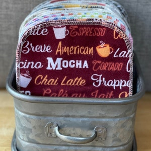 May include: A stack of red, white, and blue coffee-themed dishcloths with various coffee drink names printed on them, such as "Espresso", "Breve", "American", "Mocha", "Chai Latte", and "Frappe". The dishcloths are in a galvanized metal container with a handle.