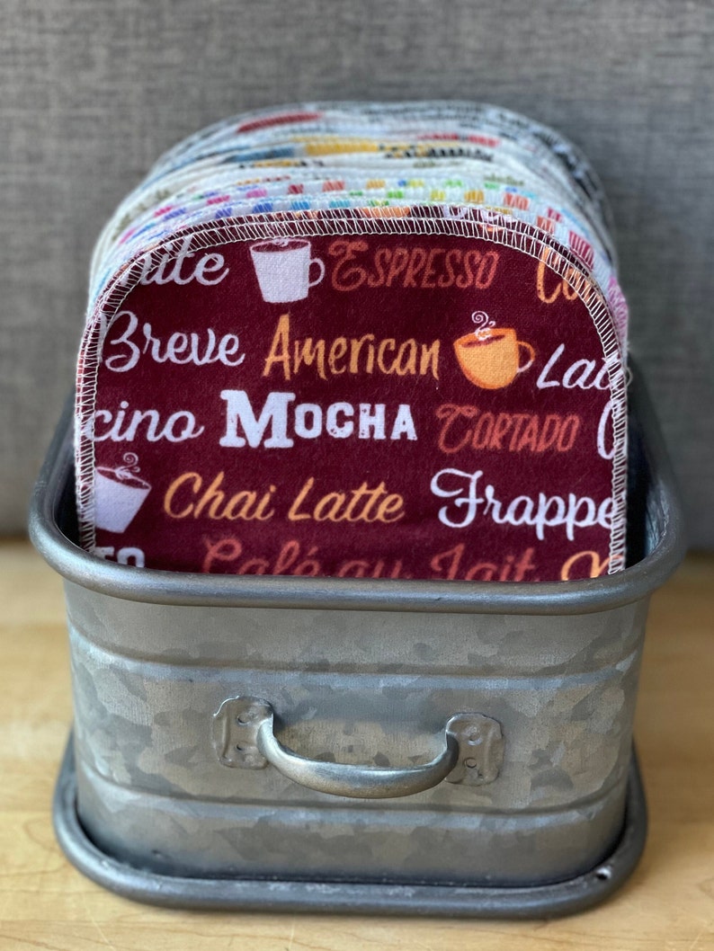 May include: A stack of red, white, and blue coffee-themed dishcloths with various coffee drink names printed on them, such as "Espresso", "Breve", "American", "Mocha", "Chai Latte", and "Frappe". The dishcloths are in a galvanized metal container with a handle.