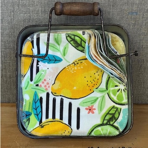 May include: A square metal holder with a dark wooden handle containing a stack of twelve square fabric cloths. The cloths feature assorted patterns including yellow lemons, green limes, black vertical stripes, and small pink flowers.