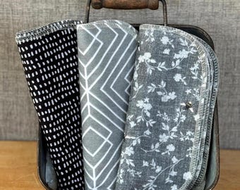 May include: A dark metal caddy with a wooden handle containing three folded fabric cloths. The cloths feature distinct patterns: a black and white dash print, a gray and white geometric line design, and a gray floral print. Text reads 4 Each pattern A.