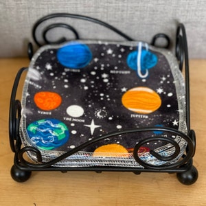 May include: A black metal napkin holder with a stack of fabric napkins featuring a space theme. The napkins have a black background with white stars and colorful planets, including Venus, Jupiter, and the Moon.