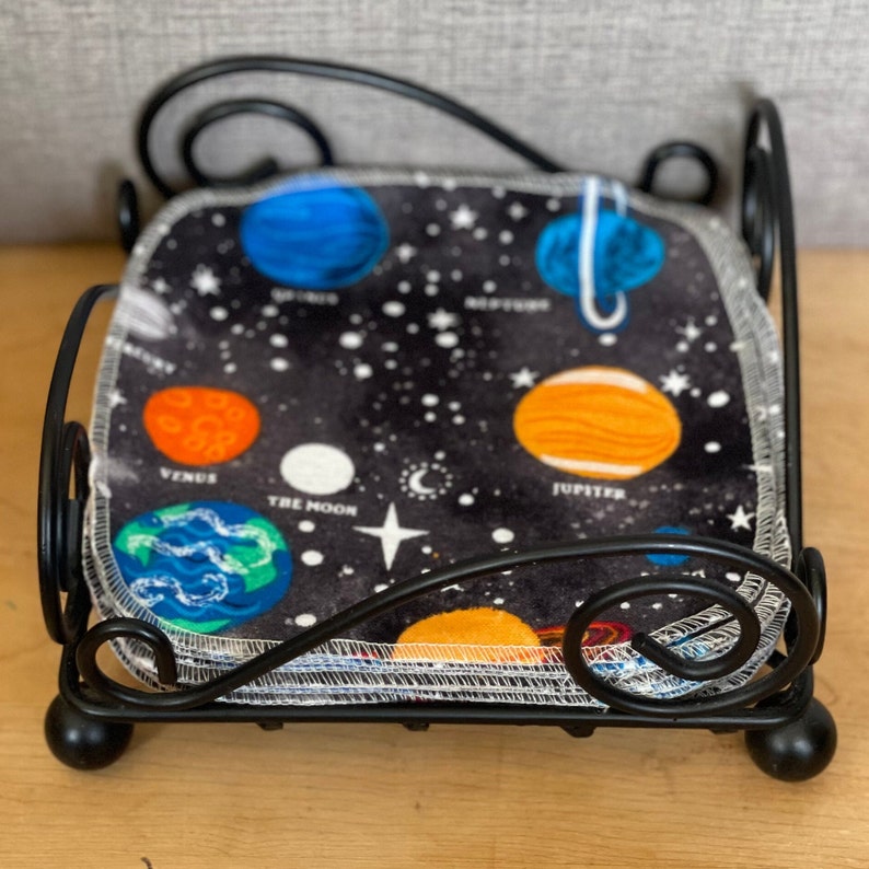 May include: A black metal napkin holder with a stack of fabric napkins featuring a space theme. The napkins have a black background with white stars and colorful planets, including Venus, Jupiter, and the Moon.