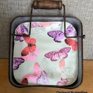 May include: A square metal basket with a dark brown wire frame and a wooden handle, containing a fabric square featuring a light green background with a pattern of butterflies in shades of pink, purple, and red. The text Orchard Butterflies is visible.