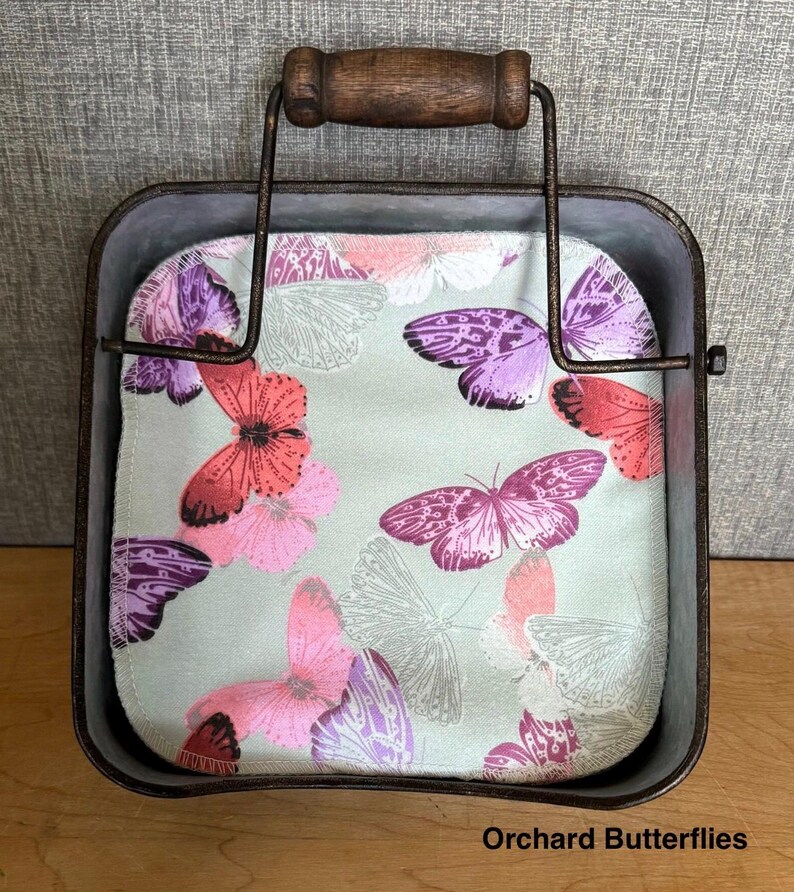 May include: A square metal basket with a dark brown wire frame and a wooden handle, containing a fabric square featuring a light green background with a pattern of butterflies in shades of pink, purple, and red. The text Orchard Butterflies is visible.