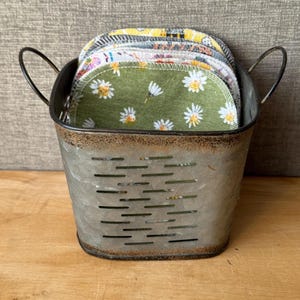 May include: A galvanized metal bucket with two handles is filled with several reusable fabric rounds. The rounds have various floral patterns, including daisies on a green background. The bucket sits on a wooden surface.