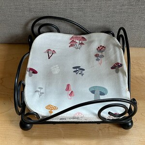 May include: Black metal napkin holder with a white fabric napkin featuring a mushroom pattern. The napkin has a white background with red, pink, blue, and green mushrooms.