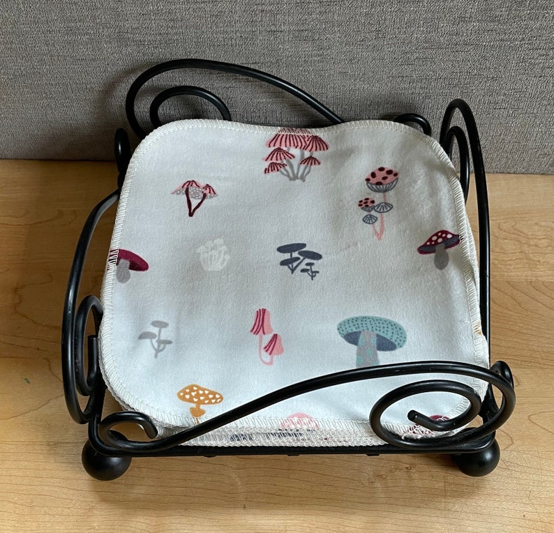 May include: Black metal napkin holder with a white fabric napkin featuring a mushroom pattern. The napkin has a white background with red, pink, blue, and green mushrooms.
