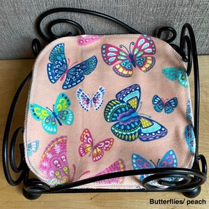 May include: A stack of square fabric cloths with a peach background and a multi-colored butterfly print, held within a black metal wire holder with decorative scrollwork. The butterflies feature shades of blue, pink, yellow, and teal.