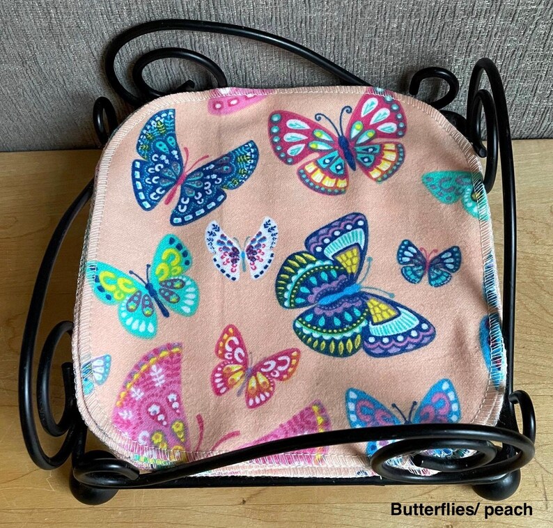 May include: A stack of square fabric cloths with a peach background and a multi-colored butterfly print, held within a black metal wire holder with decorative scrollwork. The butterflies feature shades of blue, pink, yellow, and teal.