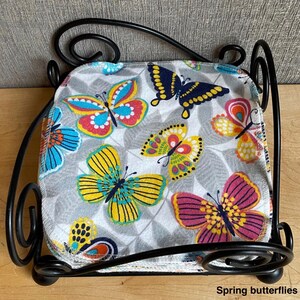 May include: A stack of square fabric cloths featuring a colorful butterfly pattern in shades of yellow, pink, blue, and orange on a gray leaf-print background, held within a black metal wire holder with decorative scrollwork and the text Spring butterflies.