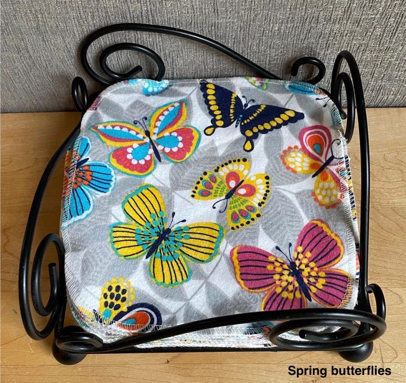 May include: A stack of square fabric cloths featuring a colorful butterfly pattern in shades of yellow, pink, blue, and orange on a gray leaf-print background, held within a black metal wire holder with decorative scrollwork and the text Spring butterflies.