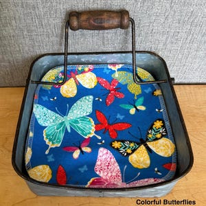 May include: A square galvanized metal caddy with a dark brown wooden handle containing a stack of fabric napkins. The napkins feature a vibrant blue background with a repeating pattern of colorful butterflies in shades of yellow, red, and teal.