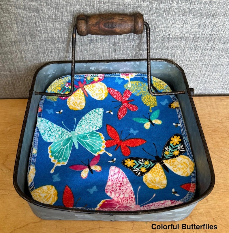 May include: A square galvanized metal caddy with a dark brown wooden handle containing a stack of fabric napkins. The napkins feature a vibrant blue background with a repeating pattern of colorful butterflies in shades of yellow, red, and teal.