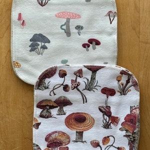 May include: Two white cotton dishcloths with a mushroom print. The top dishcloth has a light gray mushroom print with pink and red accents. The bottom dishcloth has a brown and red mushroom print.