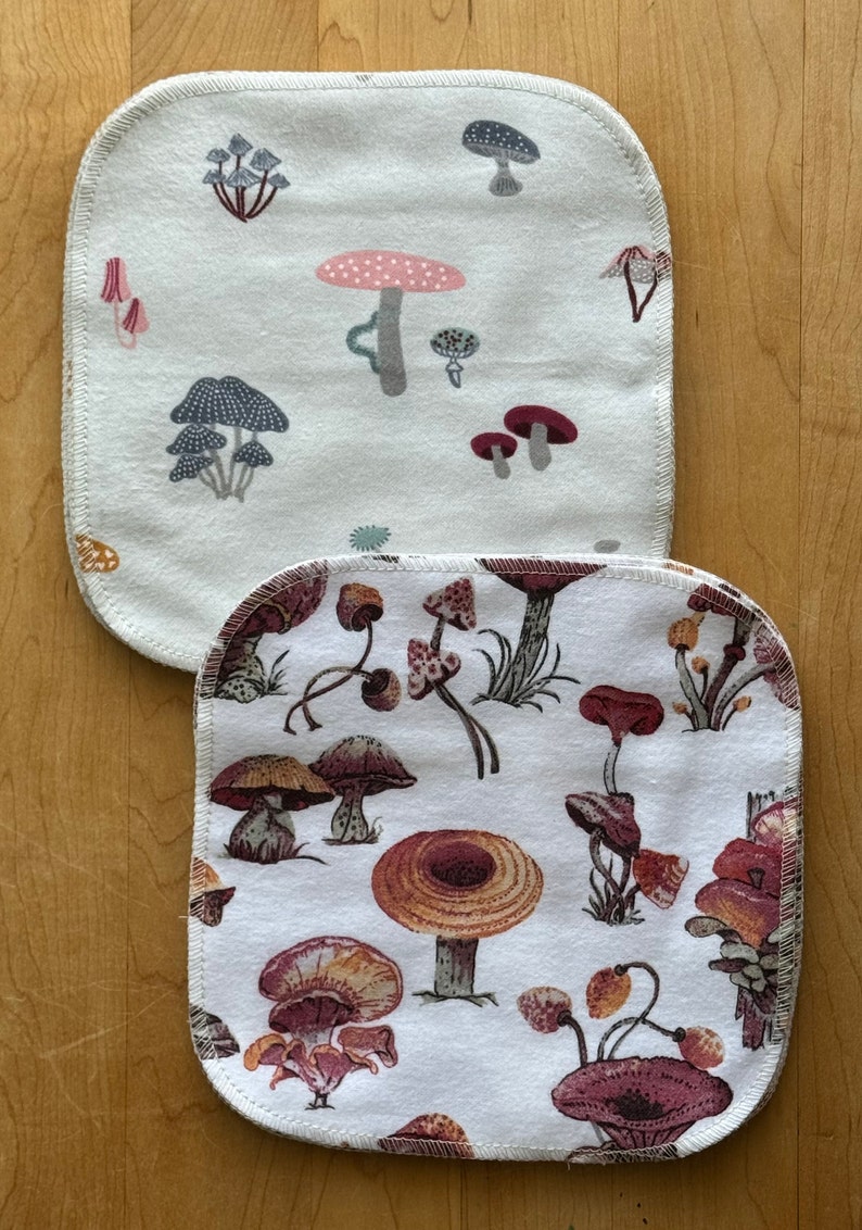 May include: Two white cotton dishcloths with a mushroom print. The top dishcloth has a light gray mushroom print with pink and red accents. The bottom dishcloth has a brown and red mushroom print.