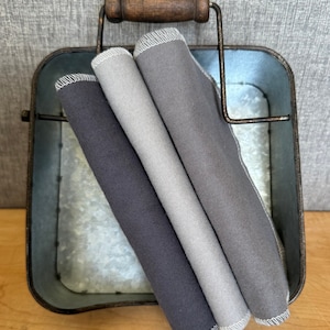 May include: Three gray fabric dishcloths folded and stacked in a metal container with a wooden handle.