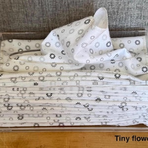 May include: A clear acrylic rectangular tissue box holder containing a stack of white fabric napkins or tissues featuring a repeating pattern of small black flowers. The text "Tiny flowers / WT" is printed in the bottom right corner.