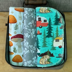 May include: Three fabric dish towels with different patterns. The first towel has a white background with orange and red mushrooms. The second towel has a gray background with white flowers. The third towel has a turquoise background with a camper van, trees, and bears.