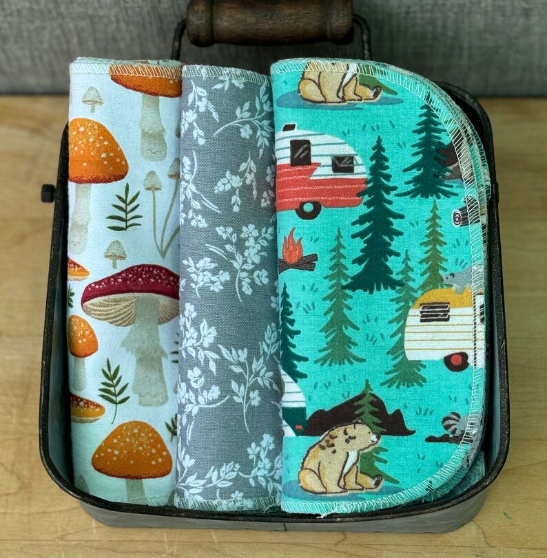 May include: Three fabric dish towels with different patterns. The first towel has a white background with orange and red mushrooms. The second towel has a gray background with white flowers. The third towel has a turquoise background with a camper van, trees, and bears.