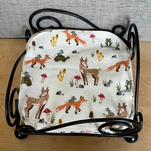 May include: A black metal doll bed with a white fabric mattress featuring a woodland animal print. The print includes deer, foxes, mice, and mushrooms.