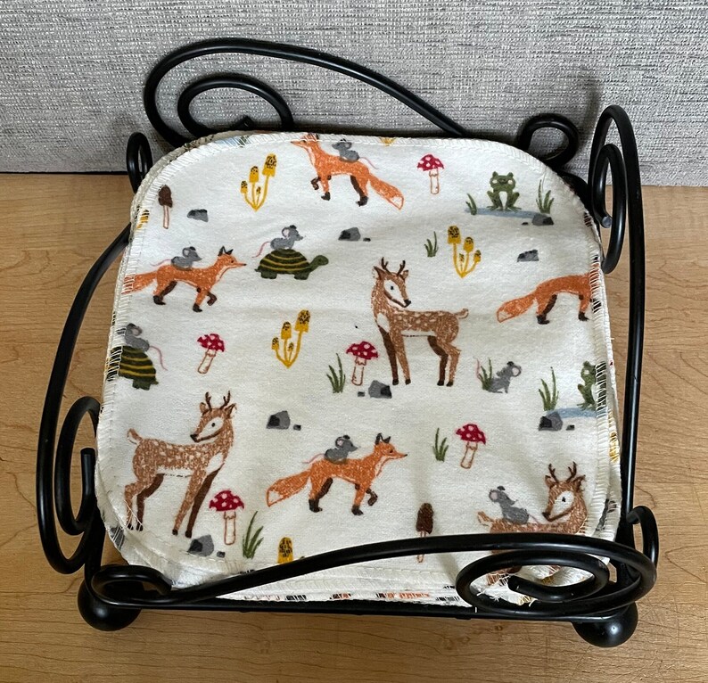 May include: A black metal doll bed with a white fabric mattress featuring a woodland animal print. The print includes deer, foxes, mice, and mushrooms.