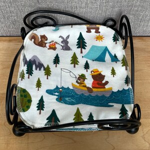 May include: A black metal napkin holder with a white fabric napkin featuring a camping theme. The napkin has illustrations of bears, squirrels, rabbits, and trees. The text on the napkin reads "Camp Out".