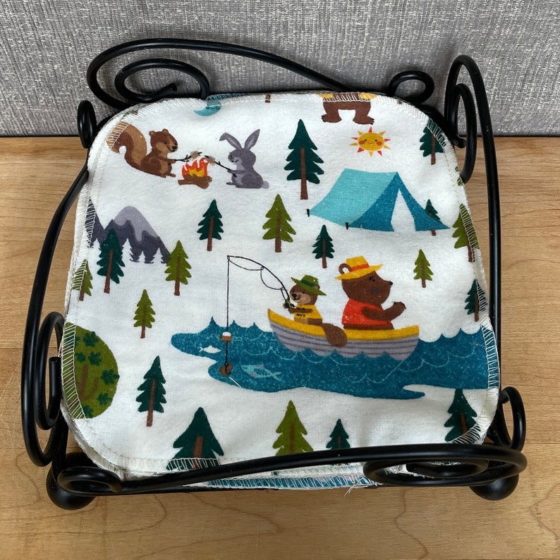 May include: A black metal napkin holder with a white fabric napkin featuring a camping theme. The napkin has illustrations of bears, squirrels, rabbits, and trees. The text on the napkin reads "Camp Out".