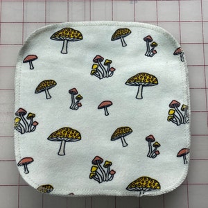 May include: A white fabric square with a repeating pattern of yellow and red mushrooms. The fabric has a white stitched border.