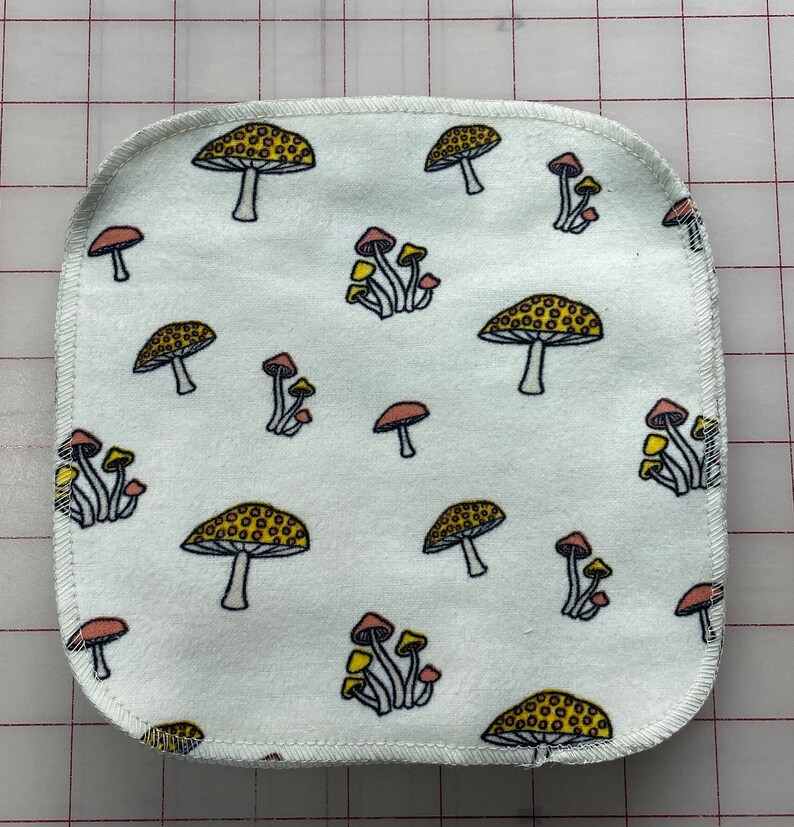 May include: A white fabric square with a repeating pattern of yellow and red mushrooms. The fabric has a white stitched border.