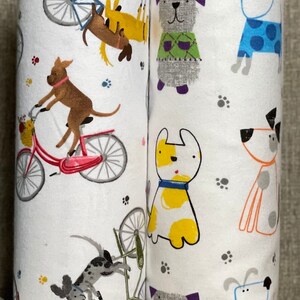 K&ouml;nnte beinhalten: Zwei wei&szlig;e Stoffrollen mit einem sich wiederholenden Muster aus Hunden und Fahrr&auml;dern. Die Hunde sind in verschiedenen Farben und Posen, darunter Fahrradfahren, Pullover tragen und mit einem Ball spielen. Die Fahrr&auml;der sind rosa und blau.