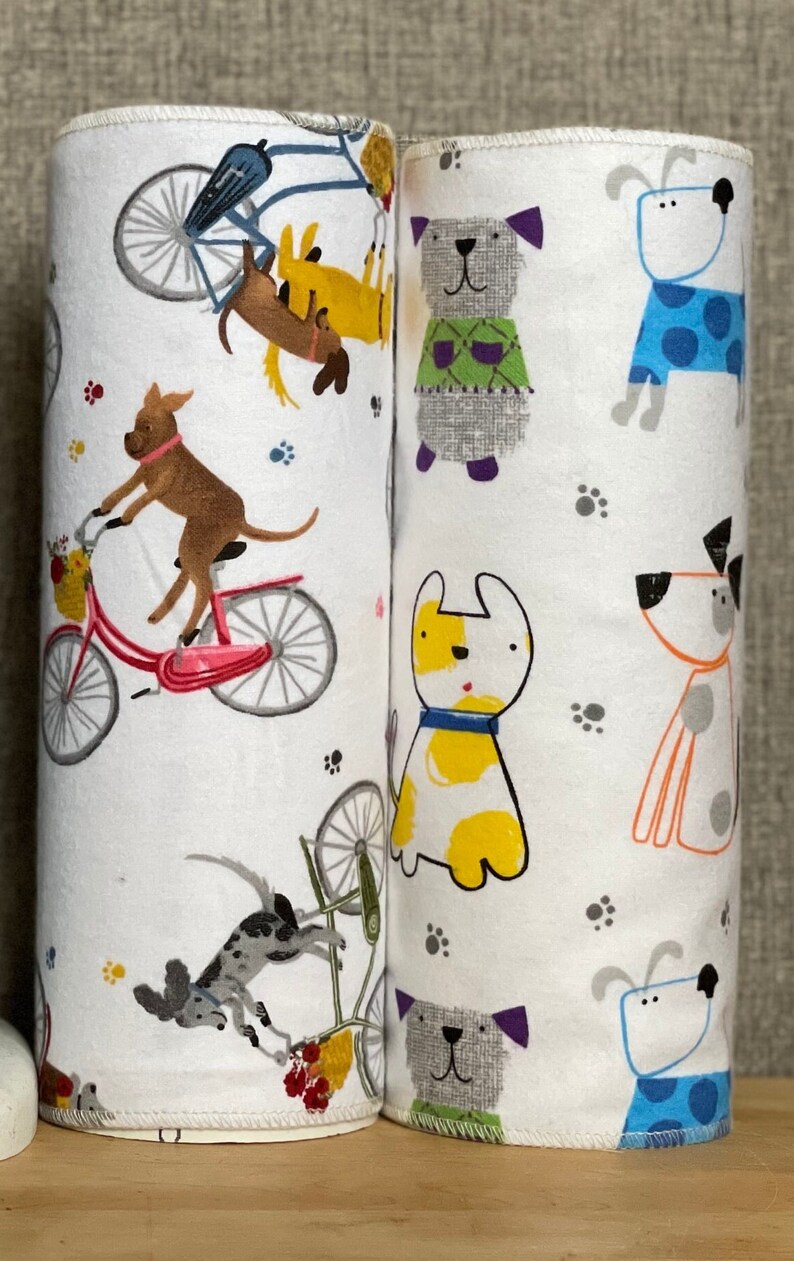 K&ouml;nnte beinhalten: Zwei wei&szlig;e Stoffrollen mit einem sich wiederholenden Muster aus Hunden und Fahrr&auml;dern. Die Hunde sind in verschiedenen Farben und Posen, darunter Fahrradfahren, Pullover tragen und mit einem Ball spielen. Die Fahrr&auml;der sind rosa und blau.