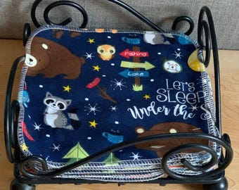 Könnte beinhalten: Ein Stapel quadratischer Stofftücher auf dunkelblauem Grund mit Zeichentrickbären, einem Waschbären, einer Eule und Campingschildern. Der Text lautet "Let's SLEEP Under the Stars". Die Tücher stecken in einem schwarzen Metalldraht-Halter.
