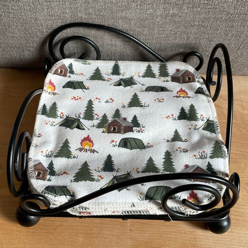 May include: Black metal napkin holder with a white fabric liner featuring a camping themed print with green trees, red cabins, orange campfires, and blue tents.