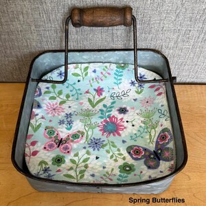 May include: A square galvanized metal napkin holder with a dark brown wooden handle. Inside sits a stack of napkins featuring a light blue background with a colorful pattern of pink and purple butterflies, green leaves, and various floral designs. Text reads Spring Butterflies.